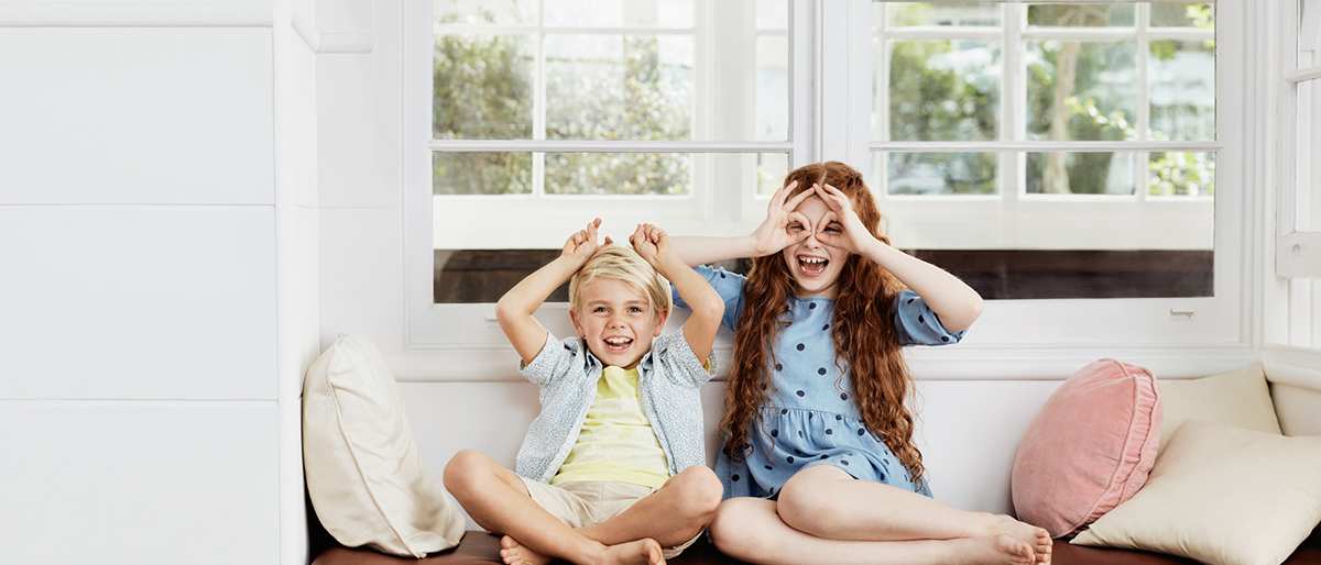 and young brother and sister sit comfortably next to eachother making horns and glasses on their faces with their hands