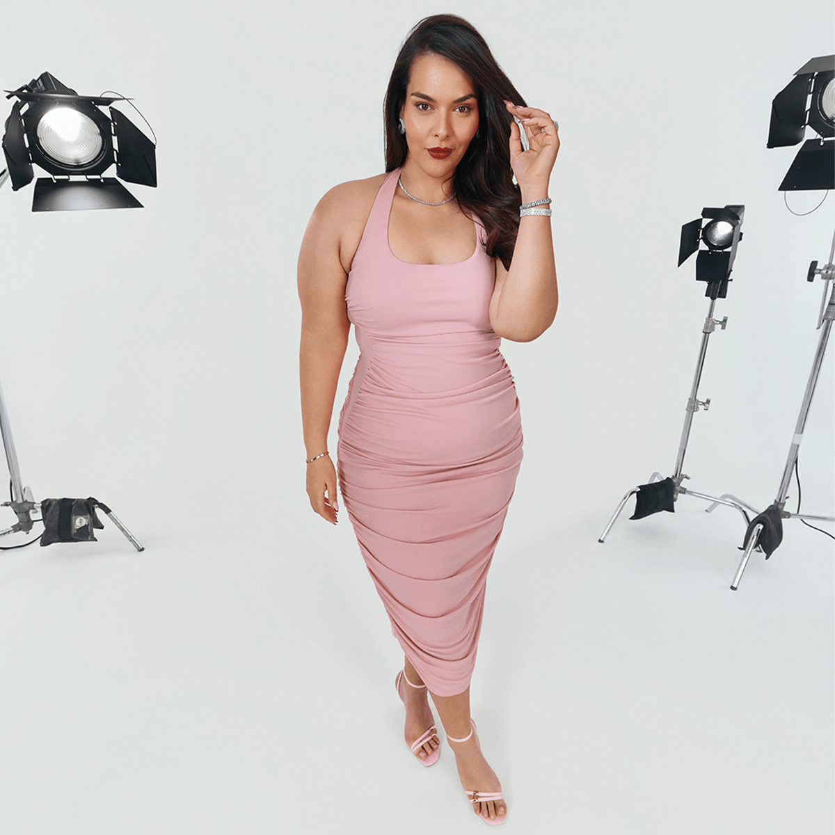 A woman wearing a pastel pink bodycon dress with pink strappy heels and silver jewellery