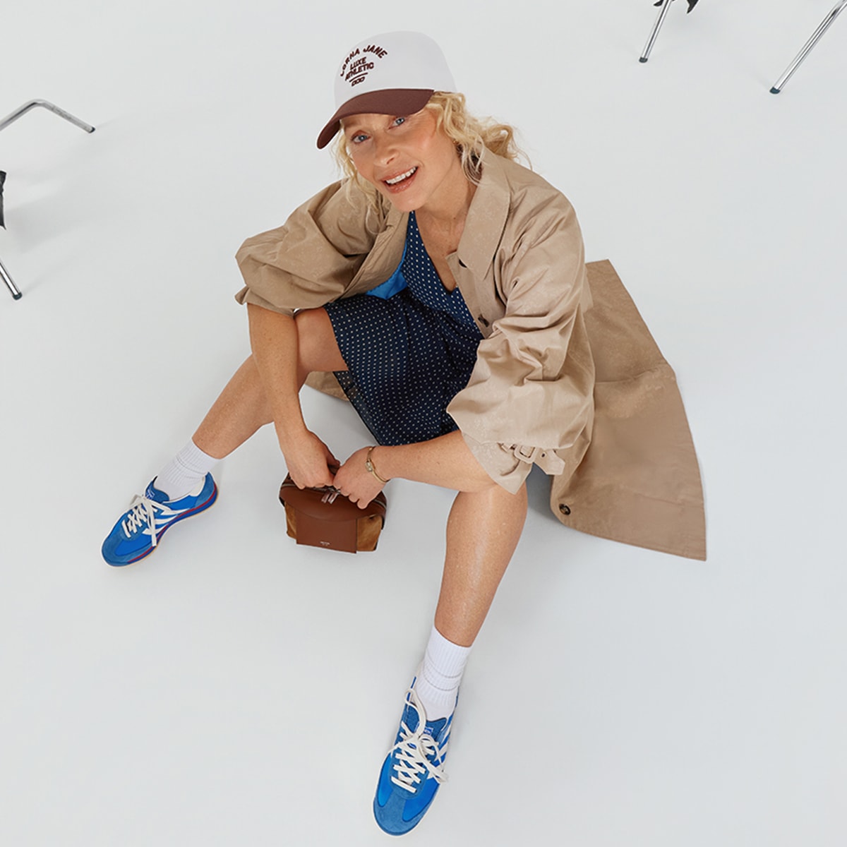 A woman sitting on the ground with bent legs, wearing a blue polka dot dress, trench coat, blue sneakers and a cap