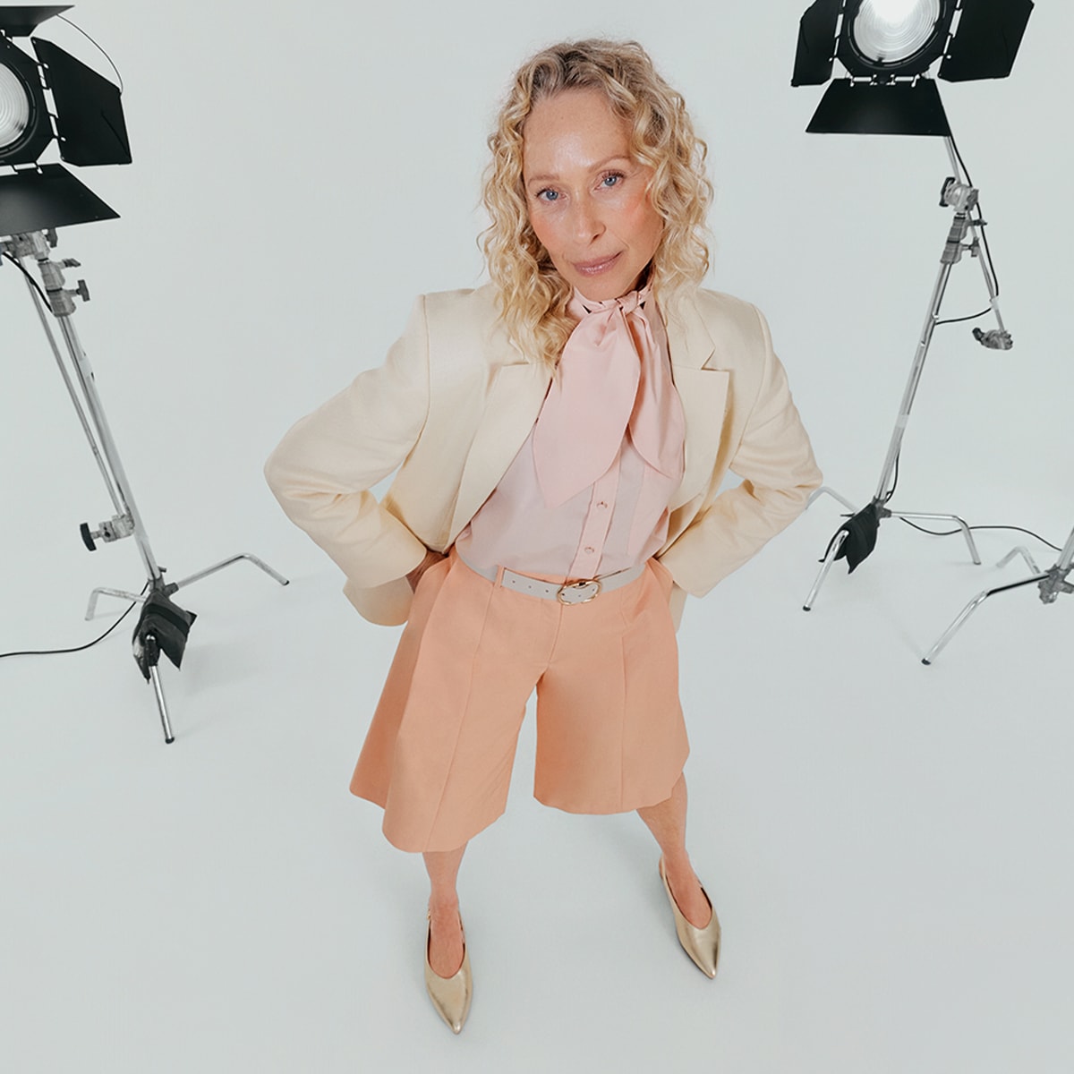 A woman standing with hands on hips, wearing apricot tailored shorts, a cream blazer and pink shirt