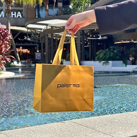 Hand holding a gold Palermo Perfumes bag, against a background of a pond in a shopping centre.